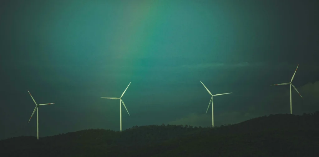 Empat turbin angin berdiri di atas perbukitan hijau gelap di bawah langit berawan dengan gradasi warna kebiruan dan sedikit pelangi samar di tengah langit.
