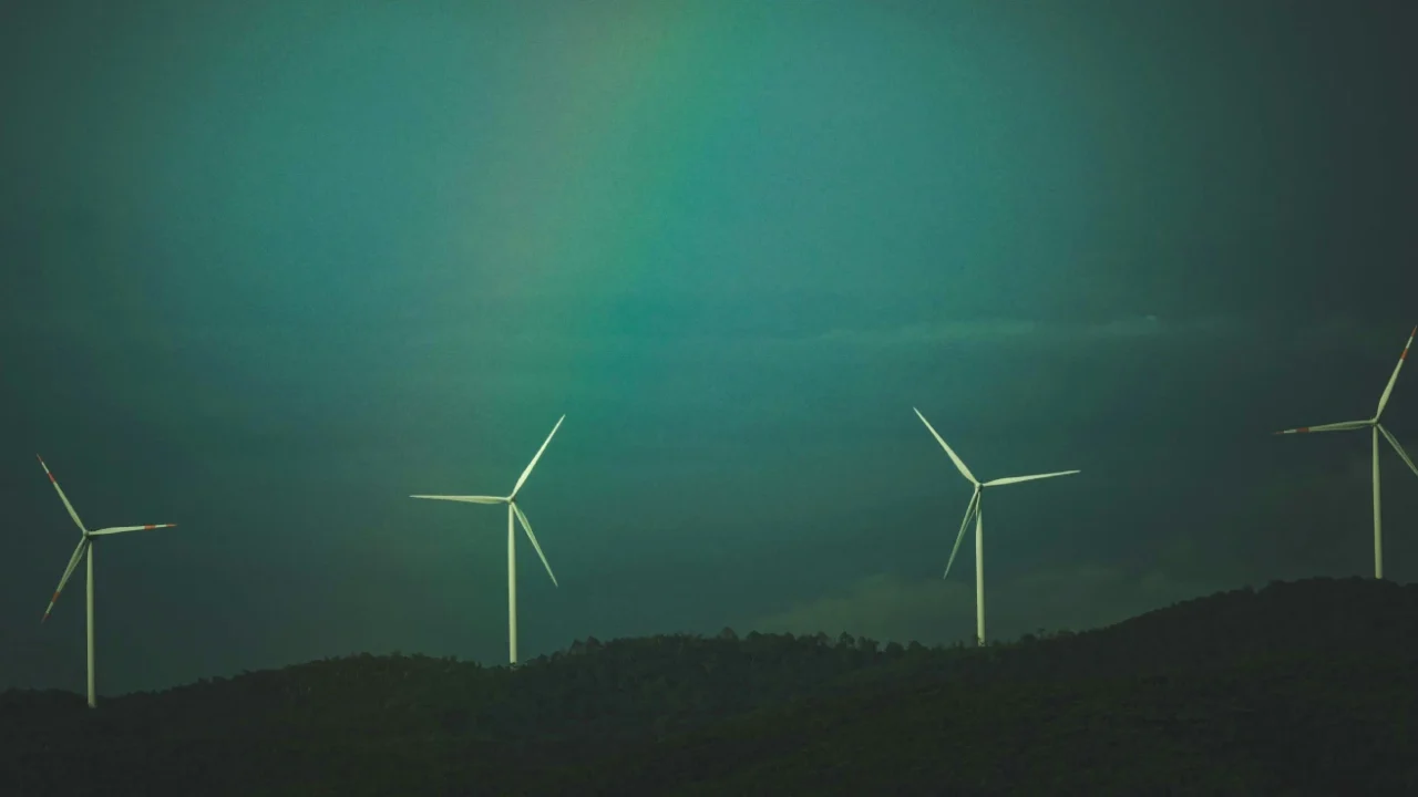 Empat turbin angin berdiri di atas perbukitan hijau gelap di bawah langit berawan dengan gradasi warna kebiruan dan sedikit pelangi samar di tengah langit.