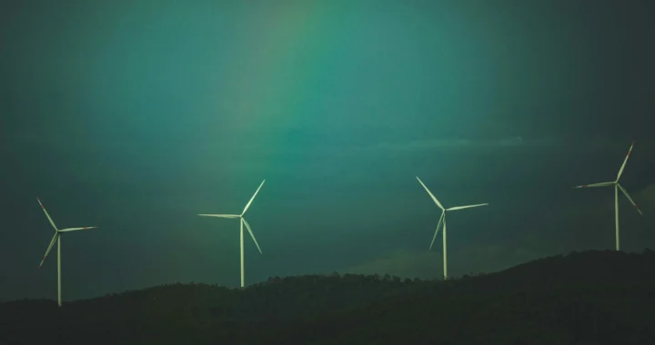 Empat turbin angin berdiri di atas perbukitan hijau gelap di bawah langit berawan dengan gradasi warna kebiruan dan sedikit pelangi samar di tengah langit.