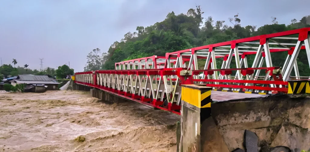 Jembatan dengan rangka baja berwarna merah dan putih yang rusak di bagian ujungnya akibat banjir besar. Air sungai keruh berwarna cokelat terlihat mengalir deras di bawahnya, dengan sebagian tanah dan aspal yang amblas di dekat jembatan.