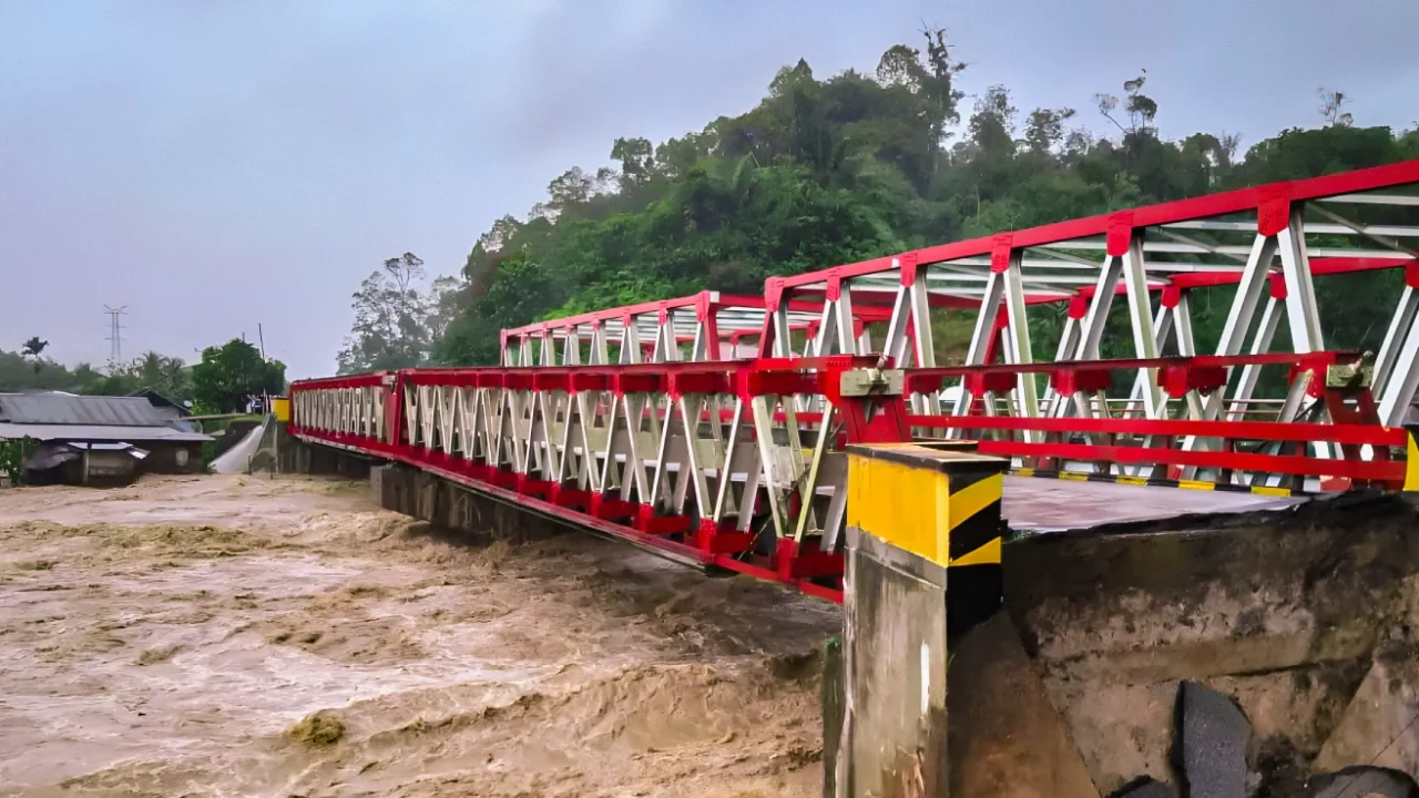 Jembatan dengan rangka baja berwarna merah dan putih yang rusak di bagian ujungnya akibat banjir besar. Air sungai keruh berwarna cokelat terlihat mengalir deras di bawahnya, dengan sebagian tanah dan aspal yang amblas di dekat jembatan.