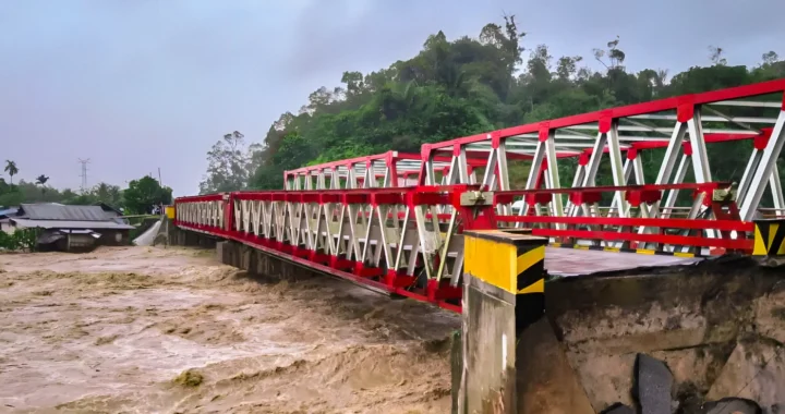 Jembatan dengan rangka baja berwarna merah dan putih yang rusak di bagian ujungnya akibat banjir besar. Air sungai keruh berwarna cokelat terlihat mengalir deras di bawahnya, dengan sebagian tanah dan aspal yang amblas di dekat jembatan.