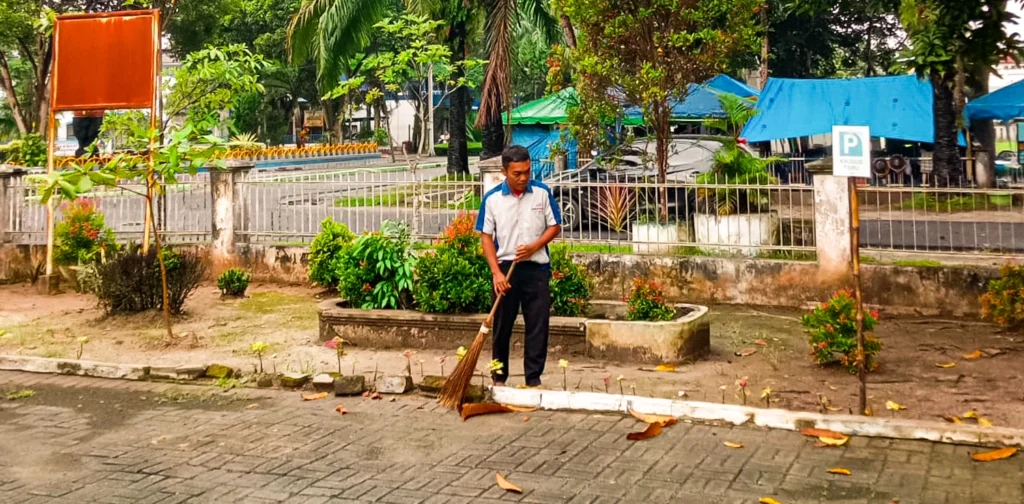 seorang pria dengan kemeja putih biru sedang menyapu dedaunan di halaman
