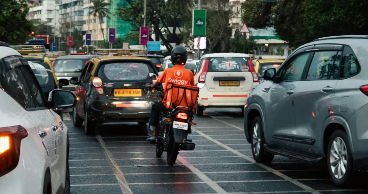 Seorang pengantar barang berjaket oranye terang melaju di tengah kemacetan lalu lintas India yang padat dengan sepeda motornya.