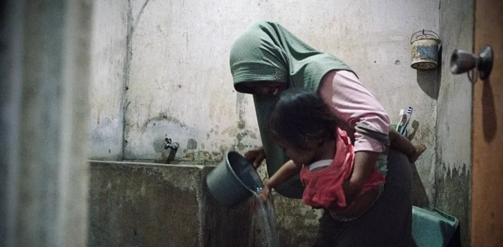 seorang perempuan menggendong anak dan mengguyurkan air dari gayung abu-abu