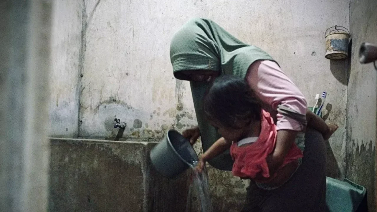 seorang perempuan menggendong anak dan mengguyurkan air dari gayung abu-abu