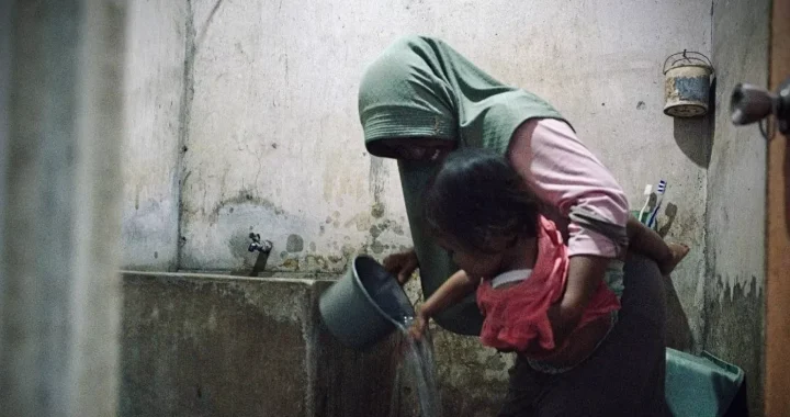 seorang perempuan menggendong anak dan mengguyurkan air dari gayung abu-abu