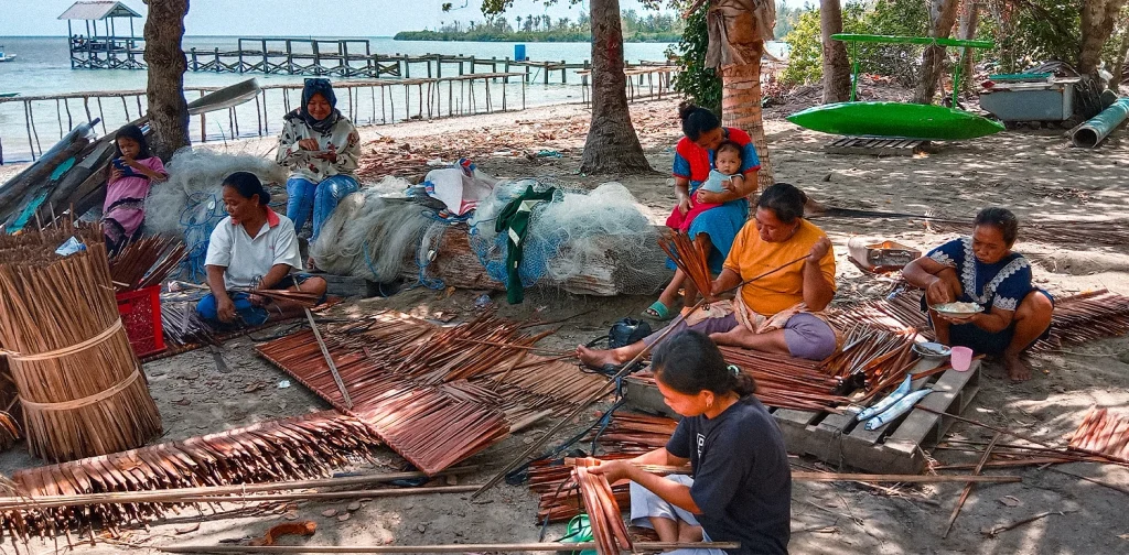 beberapa orang duduk di dekat jaring ikan di tepi pantai dan beberapa di antaranya sedang menganyam