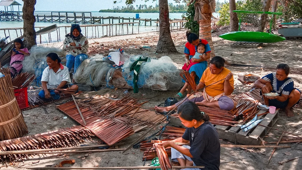 beberapa orang duduk di dekat jaring ikan di tepi pantai dan beberapa di antaranya sedang menganyam