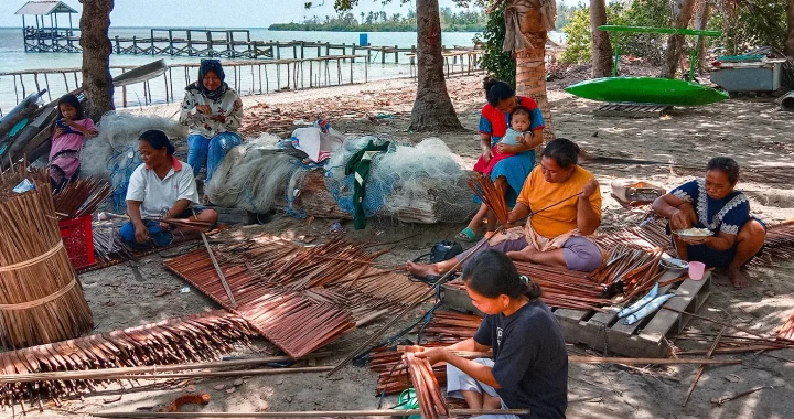 beberapa orang duduk di dekat jaring ikan di tepi pantai dan beberapa di antaranya sedang menganyam