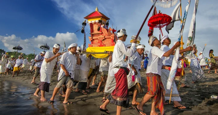 Masyarakat mengikuti prosesi upacara adat Melasti di tepi pantai, menandu perlengkapan ritual dan membawa beberapa bendera secara bersama-sama.