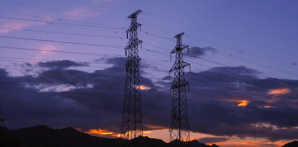 Dua menara transmisi listrik tinggi dengan latar langit senja berwarna ungu dan oranye, dengan kabel-kabel listrik membentang di atas awan gelap dan pegunungan di kejauhan.