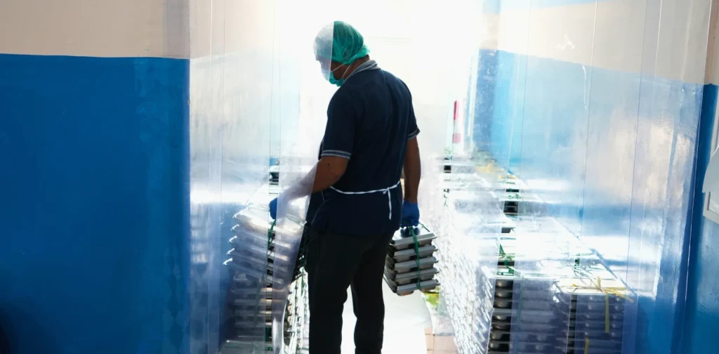 seorang petugas dengan penutup kepala dan masker menenteng tumpukan ompreng makanan berbahan baja