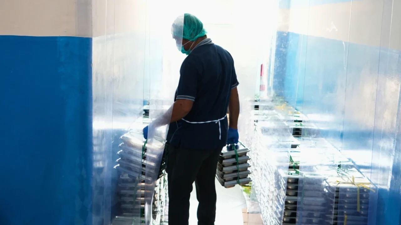 seorang petugas dengan penutup kepala dan masker menenteng tumpukan ompreng makanan berbahan baja