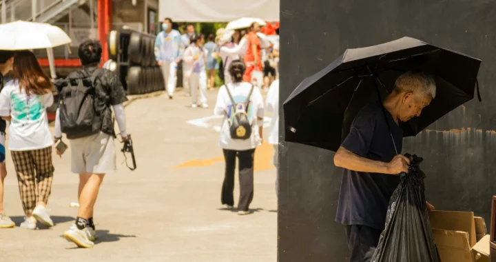 Seorang lelaki tua berdiri di pinggir jalan yang ramai dan cerah, sedang memilah-milah kardus seraya memegang payung hitam