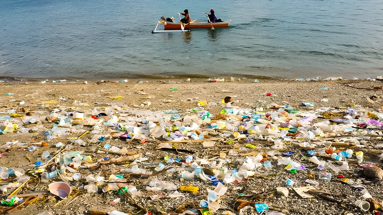 sampah plastik berserakan di tepi pantai dengan latar dua orang mendayung perahu