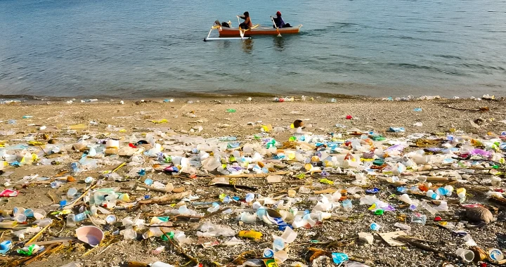 sampah plastik berserakan di tepi pantai dengan latar dua orang mendayung perahu
