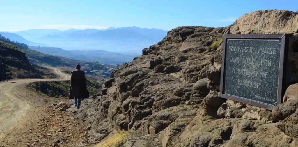 Seseorang berjalan di sepanjang jalan tanah pegunungan yang menghadap bentang alam dataran tinggi Lesotho yang luas, dengan pegunungan berlapis-lapis di kejauhan dan sebuah plakat batu bertuliskan “Wayfarer, pause and look upon a pathway to paradise.”
