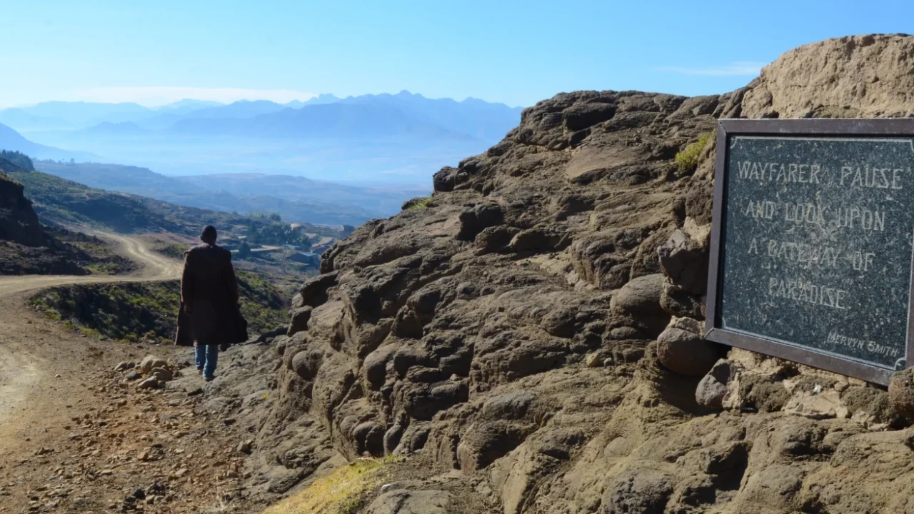 Seseorang berjalan di sepanjang jalan tanah pegunungan yang menghadap bentang alam dataran tinggi Lesotho yang luas, dengan pegunungan berlapis-lapis di kejauhan dan sebuah plakat batu bertuliskan “Wayfarer, pause and look upon a pathway to paradise.”