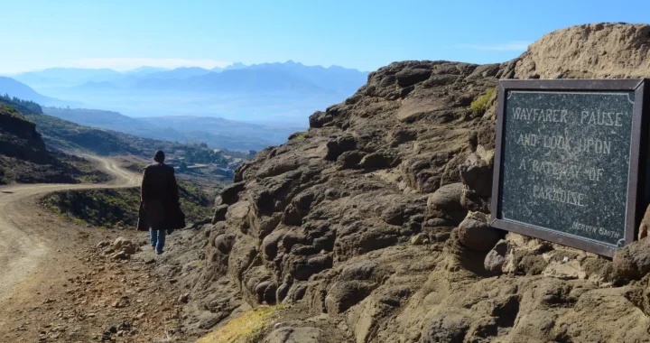Seseorang berjalan di sepanjang jalan tanah pegunungan yang menghadap bentang alam dataran tinggi Lesotho yang luas, dengan pegunungan berlapis-lapis di kejauhan dan sebuah plakat batu bertuliskan “Wayfarer, pause and look upon a pathway to paradise.”