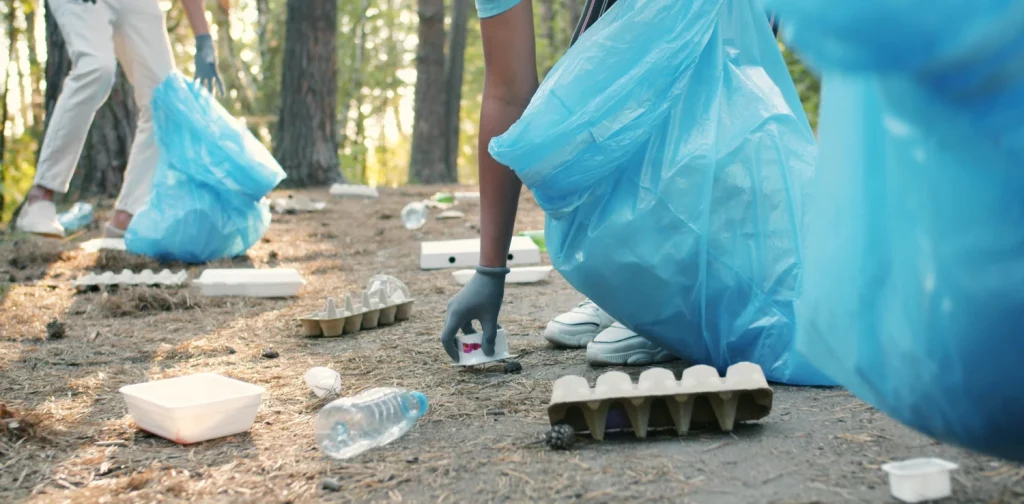 Orang-orang mengenakan sarung tangan mengumpulkan sampah ke dalam kantong sampah biru besar di area berhutan, memungut botol plastik, wadah makanan, dan kardus dari hutan.