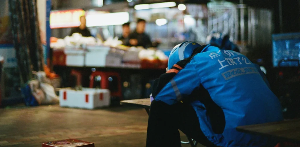 Seorang pengemudi ojek online mengenakan jaket dan helm biru Ele.me beristirahat dengan menyandarkan kepalanya di meja pasar malam, tampak kelelahan di tengah jam kerjanya.
