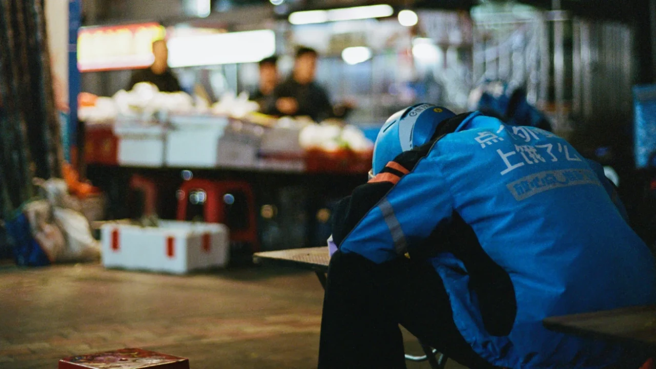 Seorang pengemudi ojek online mengenakan jaket dan helm biru Ele.me beristirahat dengan menyandarkan kepalanya di meja pasar malam, tampak kelelahan di tengah jam kerjanya.