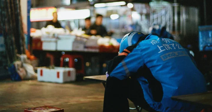 Seorang pengemudi ojek online mengenakan jaket dan helm biru Ele.me beristirahat dengan menyandarkan kepalanya di meja pasar malam, tampak kelelahan di tengah jam kerjanya.