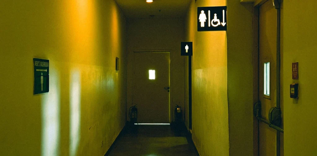 lorong dengan cahaya kuning temaram dengan tanda untuk toilet pria di bagian belakang dan tanda toilet wanita serta tanda untuk penyandang disabilitas saling berdampingan