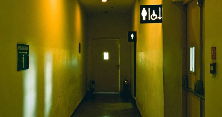 lorong dengan cahaya kuning temaram dengan tanda untuk toilet pria di bagian belakang dan tanda toilet wanita serta tanda untuk penyandang disabilitas saling berdampingan