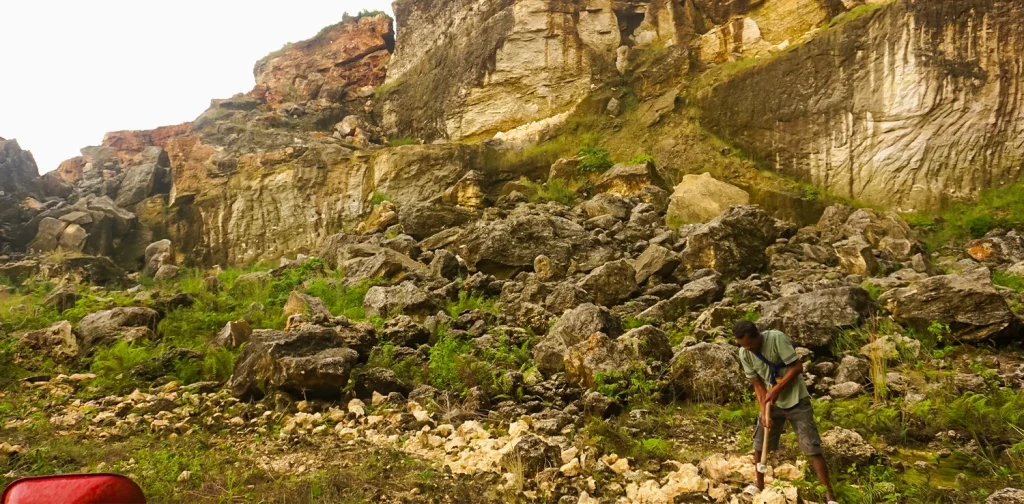 seorang pria memukul bebatuan kapur dengan palu, dengan latar bukit karst di belakangnya.
