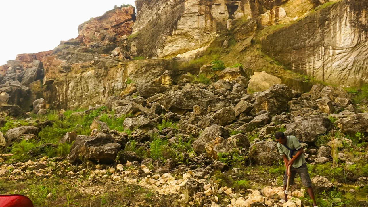 seorang pria memukul bebatuan kapur dengan palu, dengan latar bukit karst di belakangnya.