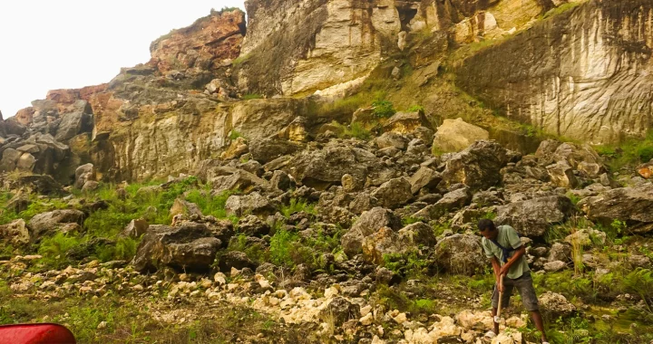 seorang pria memukul bebatuan kapur dengan palu, dengan latar bukit karst di belakangnya.