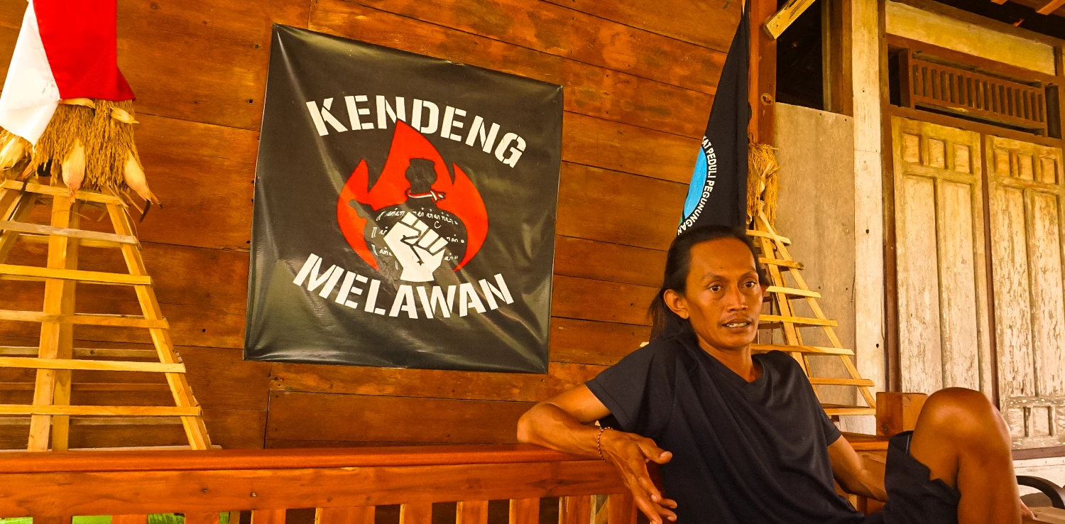 pria berkaos hitam duduk di kursi kayu dengan poster bertuliskan ‘Kendeng Melawan’ tertempel di dinging kayu di belakangnya.