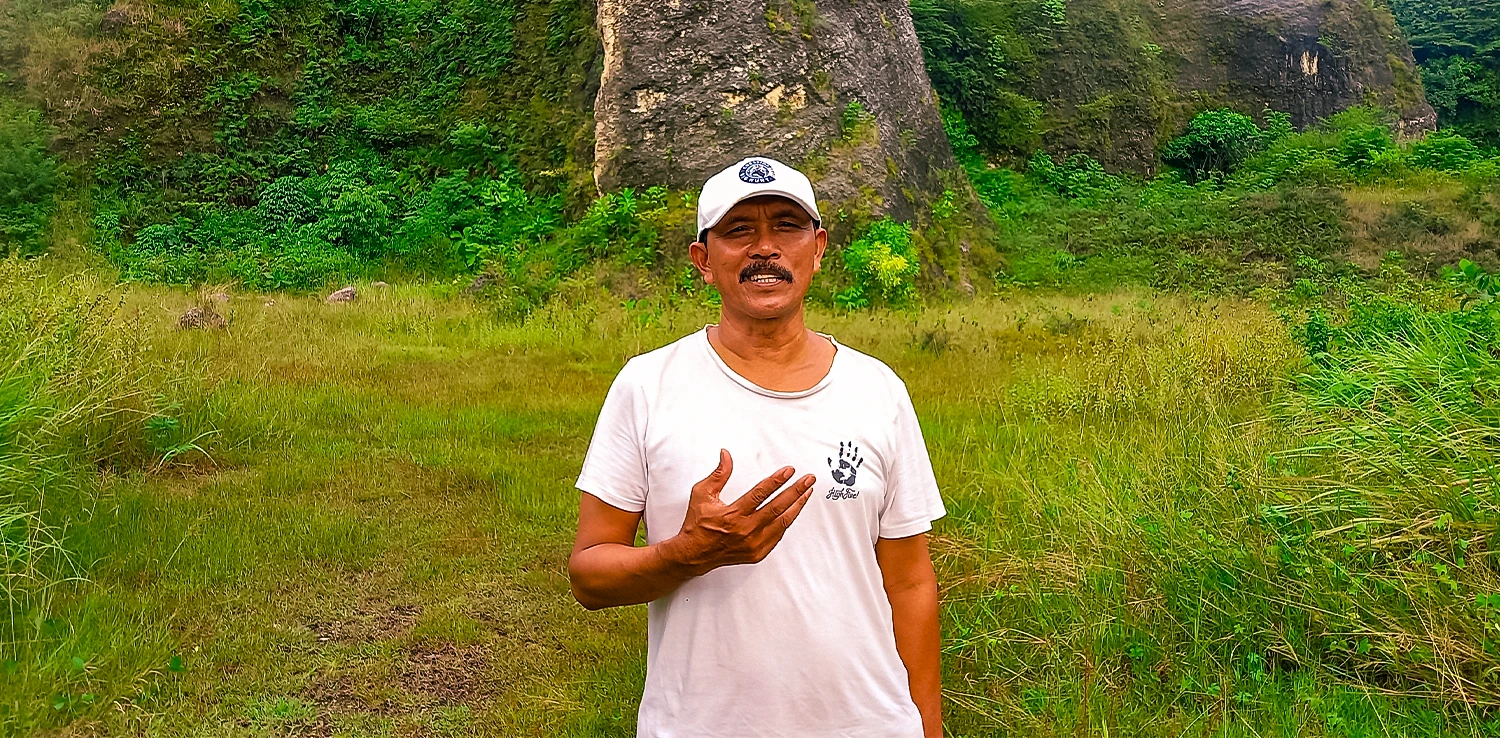 Pria bertopi putih dan berkaos putih mengangkat sebelah tangannya sambil berbicara dengan latar bukit karst.
