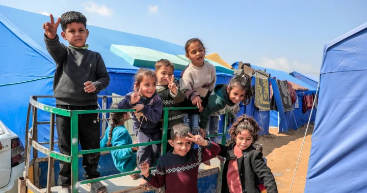 Sekelompok anak tersenyum, melambaikan tangan, dan menunjukkan tanda damai sambil berdiri di atas sebuah trailer kecil di sebuah perkemahan dengan deretan tenda biru di bawah langit yang cerah