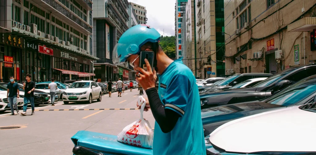 seorang pria pengantar barang berhelm dan berjaket biru sedang berdiri di samping mobil sambil berbicara di telepon