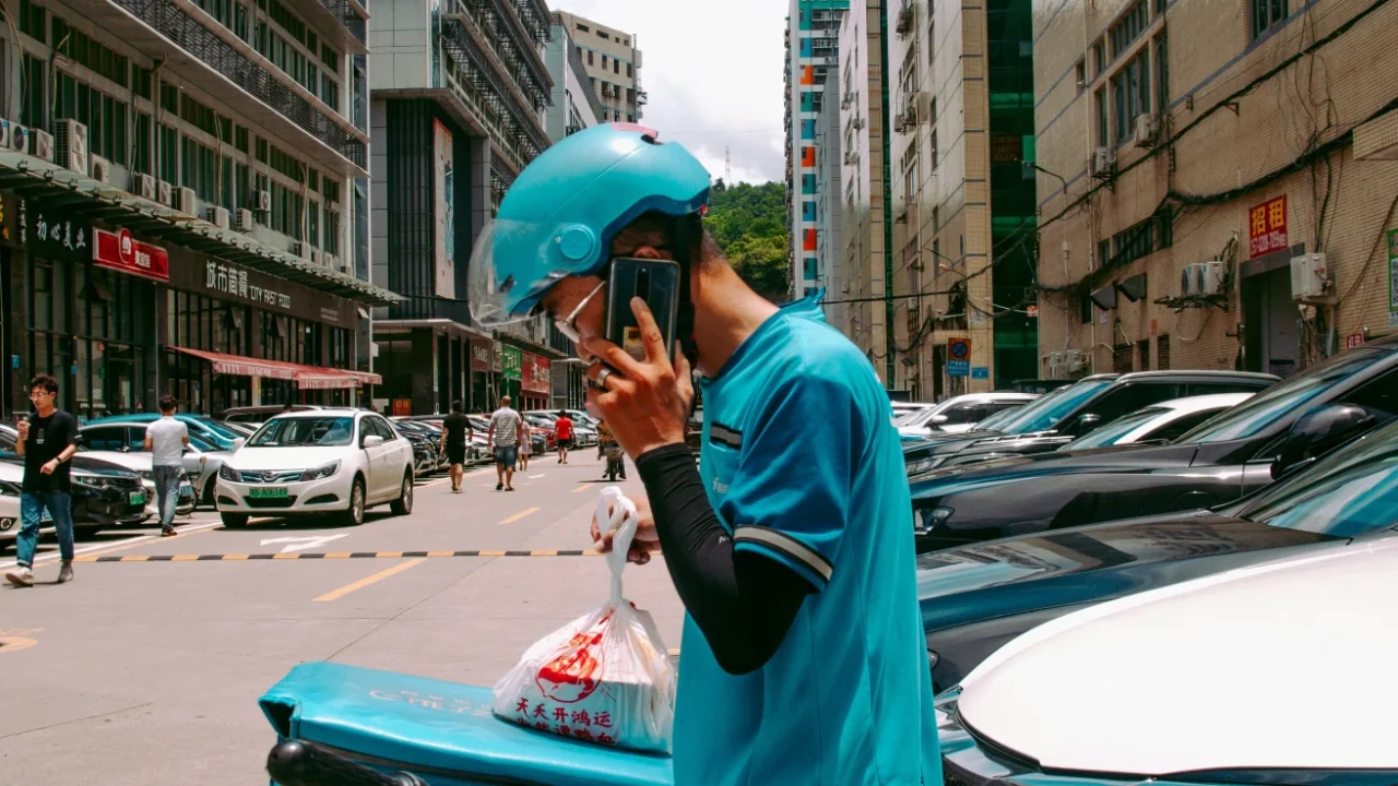 seorang pria pengantar barang berhelm dan berjaket biru sedang berdiri di samping mobil sambil berbicara di telepon