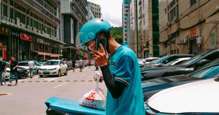 seorang pria pengantar barang berhelm dan berjaket biru sedang berdiri di samping mobil sambil berbicara di telepon