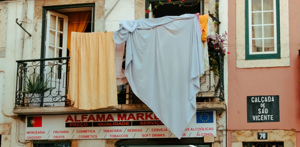 Seprai berwarna oranye dan putih dijemur menggantung dari jendela lantai dua sebuah toko di pasar lokal di Alfama, Lisbon.