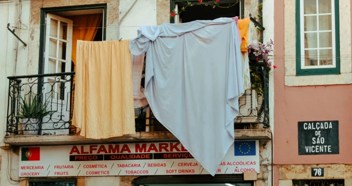 Seprai berwarna oranye dan putih dijemur menggantung dari jendela lantai dua sebuah toko di pasar lokal di Alfama, Lisbon.